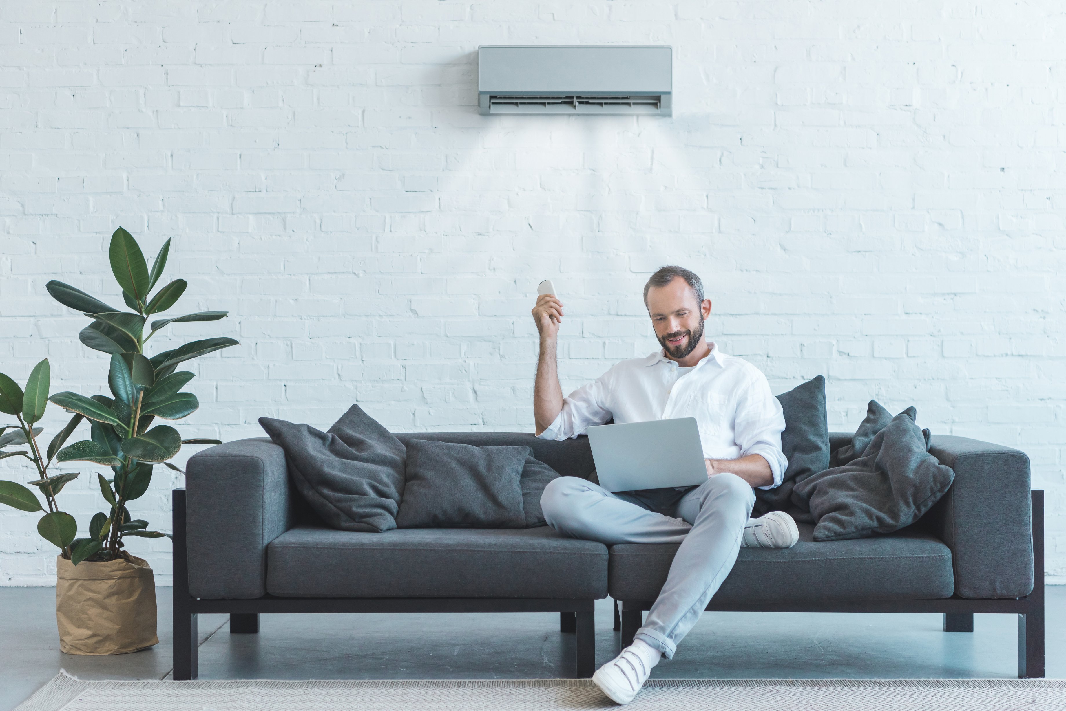 handsome man turning on air conditioner with remote control while using laptop on sofa at home